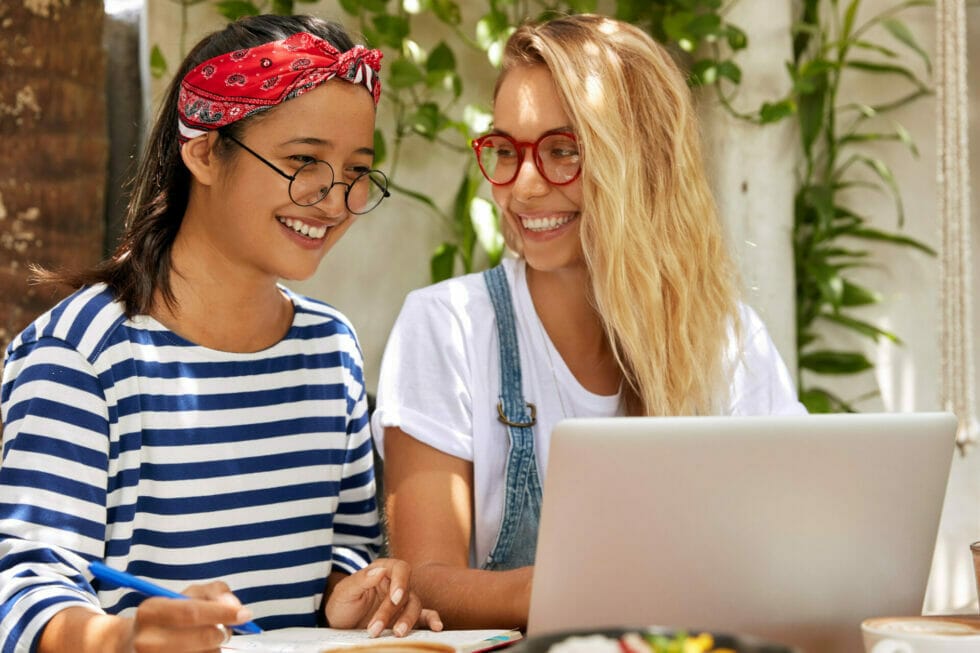 Female freelance partners get good feedback on website, look joyfully at screen of laptop computer, write information to improve internet page, pose in outdoor cozy cafeteria. Partnership concept