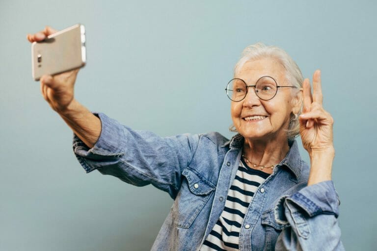 Positive smiling elderly woman wearing casual clothes and glasses poses for selfie holding smartphone in one hand and making peace or victory sign with another